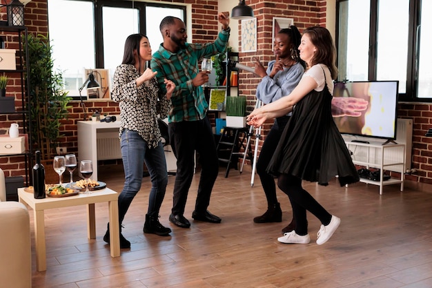 Amis joyeux ayant un événement de célébration pour l'anniversaire de l'amitié tout en buvant des boissons alcoolisées. Groupe diversifié de jeunes dansant dans le salon à la fête du vin tout en célébrant l'anniversaire.