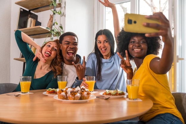 Amis multiethniques lors d'un petit-déjeuner avec jus d'orange et muffins à la maison prenant un selfie