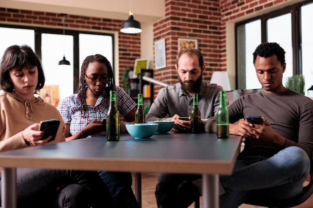 Amis multiraciaux assis à table dans le salon utilisant des smartphones tout en surfant sur Internet. Groupe diversifié de personnes assises à la maison tout en utilisant des téléphones portables modernes pour regarder du contenu vidéo.