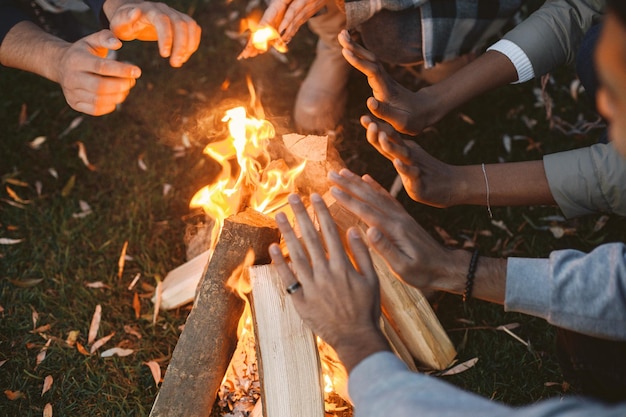 Amis multiraciaux ayant un pique-nique hors de la ville. Ils ont fait un feu et assis autour de se réchauffer les mains