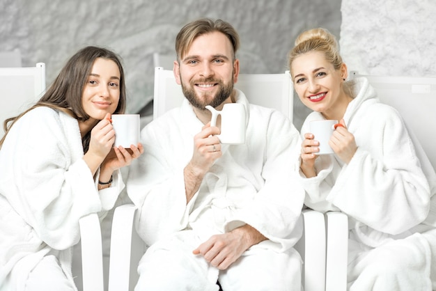 Amis en peignoir se relaxant avec des tasses à thé assis dans la salle de sel. Appliquer la thérapie par le sel au Spa