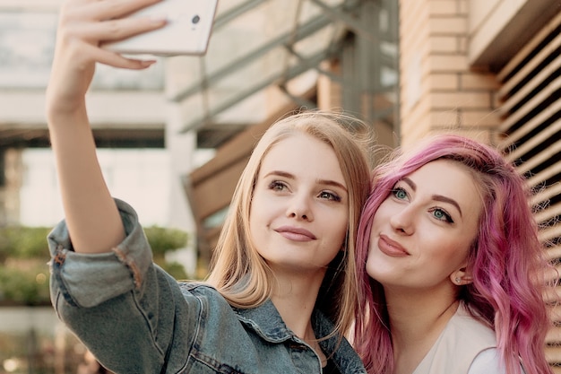 Amis prenant un selfie avec un téléphone intelligent et faisant des grimaces et s'amusant. Gros plan des filles faisant des grimaces et souriant pour selfie. Deux copines heureuses prenant une photo avec leur smartphone dans la ville