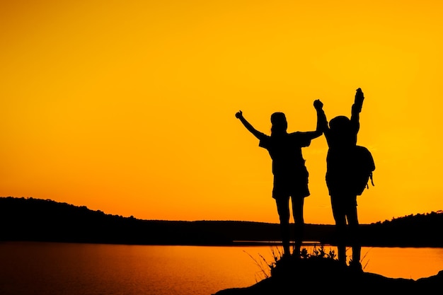 Photo des amis en silhouette debout avec les bras levés au bord du lac contre un ciel orange