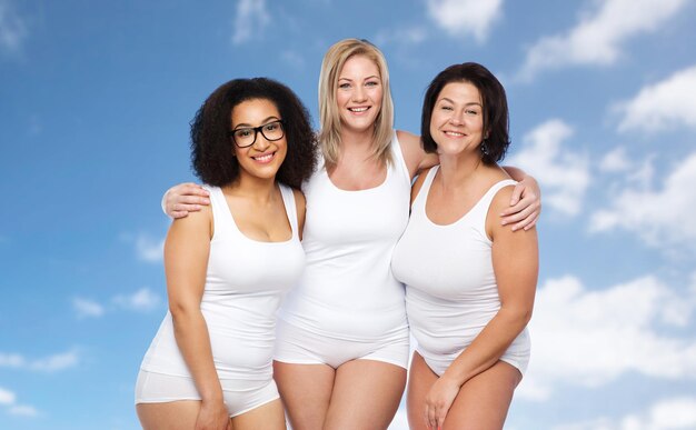 Amitié, beauté, corps positif et concept de personnes - groupe de femmes heureuses de taille plus en sous-vêtements blancs sur fond bleu ciel et nuages