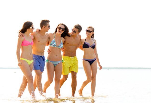 Amitié, mer, vacances d'été, vacances et concept de personnes - groupe d'amis souriants portant des maillots de bain et des lunettes de soleil marchant sur la plage