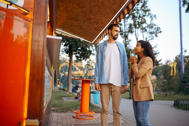 Amour couple près d'un café dans le parc d'attractions de la ville, attraction de montagnes russes. L'homme et la femme se détendent à l'extérieur. Loisirs en famille en été, thème du divertissement