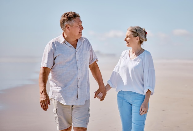 Amour à la plage et couple de personnes âgées se tenant la main en marchant pour faire de l'exercice et du bien-être