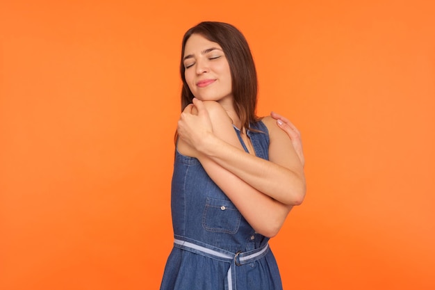 L'amour-propre et l'égoïsme. Belle femme brune narcissique en robe denim s'embrassant, se sentant en sécurité et à l'aise, satisfaite de l'apparence. studio d'intérieur tourné isolé sur fond orange