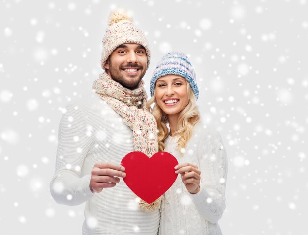 amour, saint valentin, couple, noël et concept de personnes - homme et femme souriants en chapeaux d'hiver et écharpe tenant une forme de coeur en papier rouge