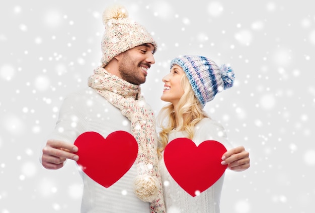 amour, saint valentin, couple, noël et concept de personnes - homme et femme souriants en chapeaux d'hiver et écharpe tenant des formes de coeur en papier rouge