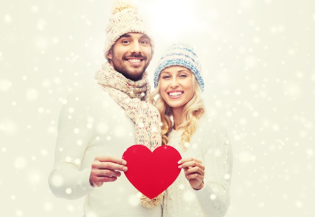 l'amour, la Saint-Valentin, le couple, Noël et les gens concept - homme et femme souriants dans des chapeaux d'hiver et une écharpe tenant une forme de cœur en papier rouge