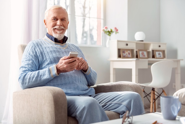 Amoureux Des Médias Sociaux. Charmant Homme âgé Optimiste Assis Dans Le Fauteuil Et Faisant Défiler La Chronologie De Ses Médias Sociaux En Souriant