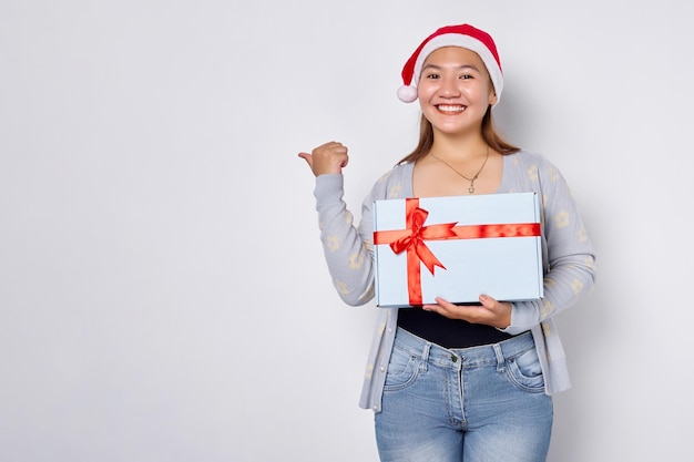 Amusant femme tenant à la main un cadeau de Noël Belle femme asiatique en chapeau de père Noël pointant les pouces de côté isolé sur fond blanc de studio Joyeux Noël Concept