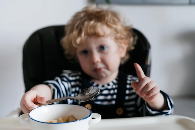 Amusant petit garçon bouclé assis sur une chaise dans la cuisine et mangeant une soupe utile