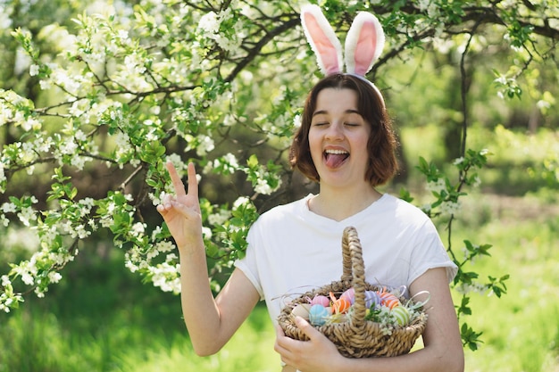 Amusante adolescente avec panier d'oeufs et oreilles de lapin sur la chasse aux oeufs de pâques dans le jardin de printemps ensoleillé