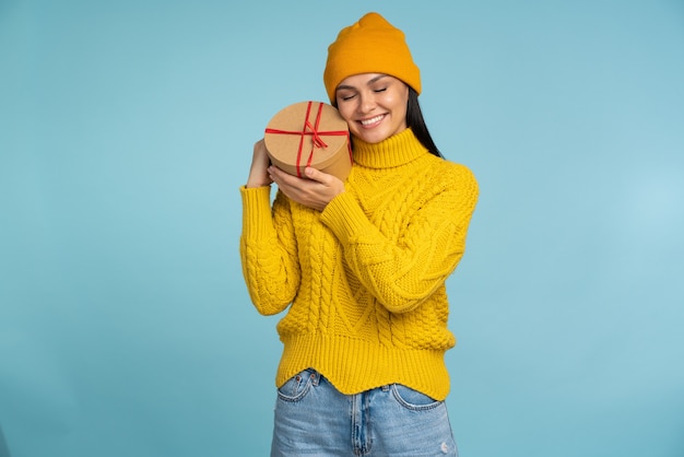 Amusante femme heureuse et heureuse embrassant une boîte-cadeau et fermant les yeux de plaisir. Personne de sexe féminin bénéficiant d'un cadeau d'anniversaire tant attendu, les rêves deviennent réalité. Studio tourné fond bleu