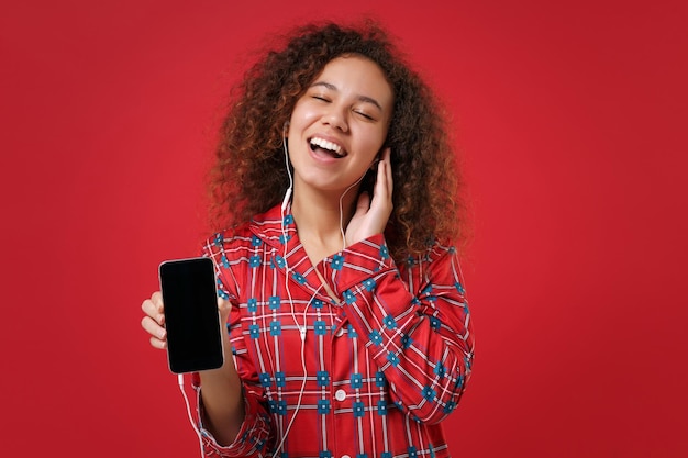 Amusante fille afro-américaine en pyjama homewear reposant à la maison isolée sur fond rouge. Détendez-vous le concept de mode de vie de bonne humeur. Écoutez de la musique avec des écouteurs, maintenez un téléphone portable avec un écran vide vide.