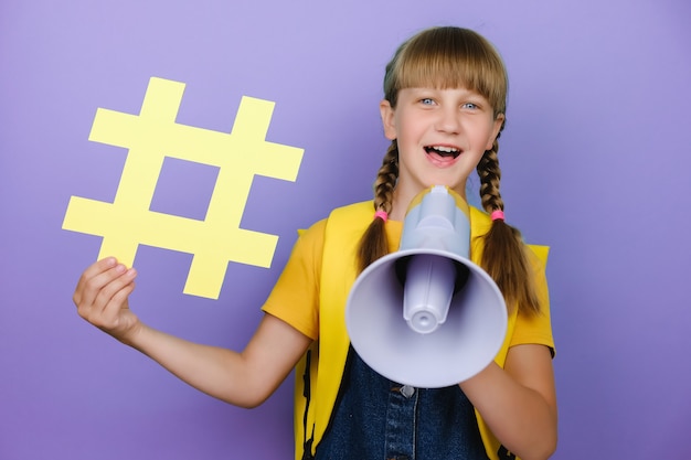 Amusante fille blonde de l'école adolescente avec un sac à dos jaune tenant un grand symbole de hashtag crier dans un mégaphone, posant isolée sur un mur de fond de couleur pourpre, un studio pour enfants. Concept de mode de vie de l'éducation