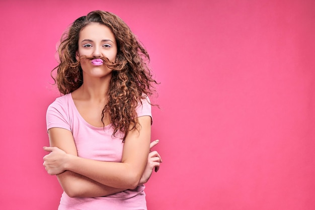 Amusante jeune femme faisant une moustache avec une mèche de cheveux isolée sur un espace de copie rose