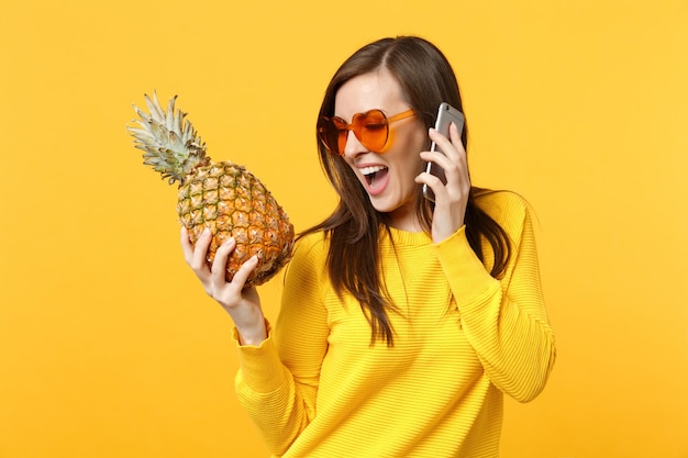 Amusante jeune femme en lunettes de soleil coeur tenant des fruits d'ananas mûrs, parlant sur un téléphone portable isolé sur fond orange jaune. Mode de vie vivant des gens, concept de vacances relaxantes. Maquette de l'espace de copie.