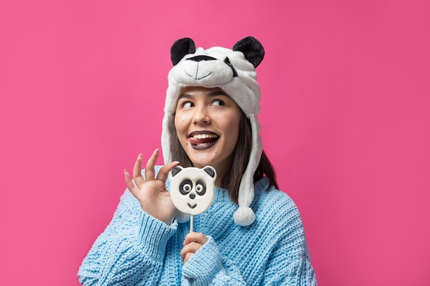 Amusante jeune fille debout avec une délicieuse sucette panda à la main et un chapeau sur la tête sur fond rose.