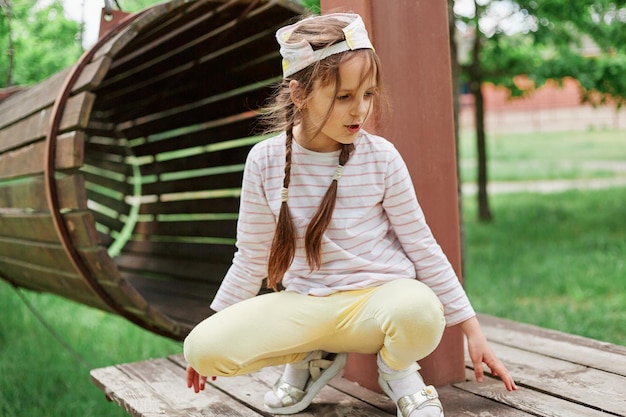 Amusante petite fille aux cheveux ébouriffés portant des vêtements décontractés et une casquette de baseball assise accroupie sur une aire de jeux près d'un tunnel rond s'amusant en plein air