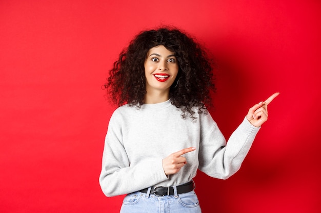 Amusé jeune femme avec une coiffure frisée, pointer du doigt droit au logo, à impressionné et excité, vérification de la promotion, mur rouge