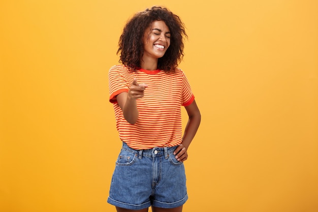 Amusée femme à la peau sombre avec une coiffure afro riant les yeux fermés indiquant joyeusement à la caméra.