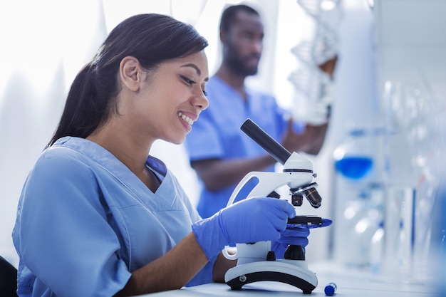 Analyse professionnelle. Joyeuse femme intelligente agréable assise devant le microscope et l'utilisant tout en étudiant des échantillons de test