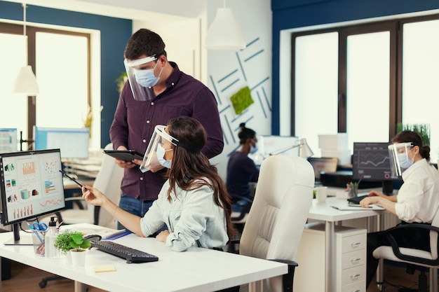 Les analystes de l'entreprise s'entraident pour terminer un projet sur le lieu de travail portant un masque facial par mesure de sécurité pour covid19. Équipe multiethnique dans un nouveau bureau financier normal dans une entreprise en tapant sur