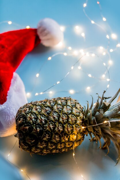 Ananas frais avec guirlandes lumineuses et chapeau de Noël sur une surface bleue.