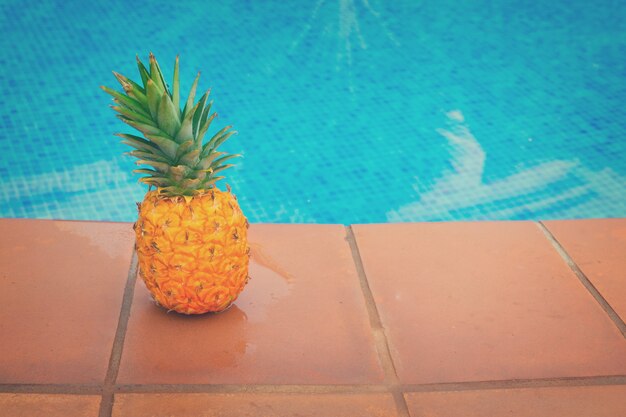 Ananas jaune entier cru et piscine carrelée avec de l'eau bleue, aux tons rétro