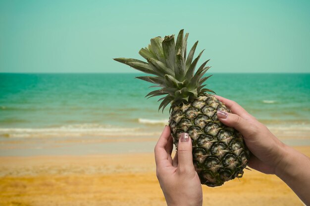 Ananas mûr sur la main de la femme avec une plage tropicale en arrière-plan Concept d'été tropical