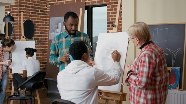 Photo ancien professeur et étudiant aidant un homme afro-américain à dessiner un vase sur toile. équipe diversifiée de personnes expliquant la technique artistique pour développer des compétences en dessin dans les cours d'art, résolutions du nouvel an.