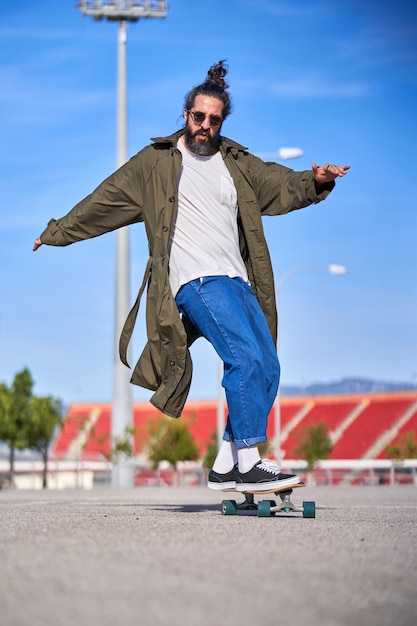 L'angle bas du corps entier a ravi l'homme en manteau et jeans tendance portant des lunettes de soleil faisant des cascades sur un longboard dans le parc par temps ensoleillé