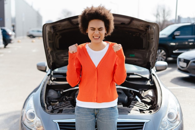 Photo angry authentic african american woman with curly hair standing by open hood of broken car