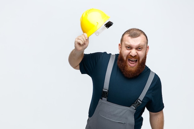 Angry young male construction worker wearing uniform lifting casque de sécurité en regardant la caméra crier isolé sur fond blanc avec copie espace