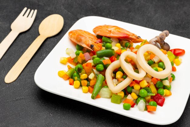 Anneaux de crevettes et de calmars avec légumes mélangés sur une assiette blanche Cuillère et fourchette sur la table Vue de dessus Fond noir