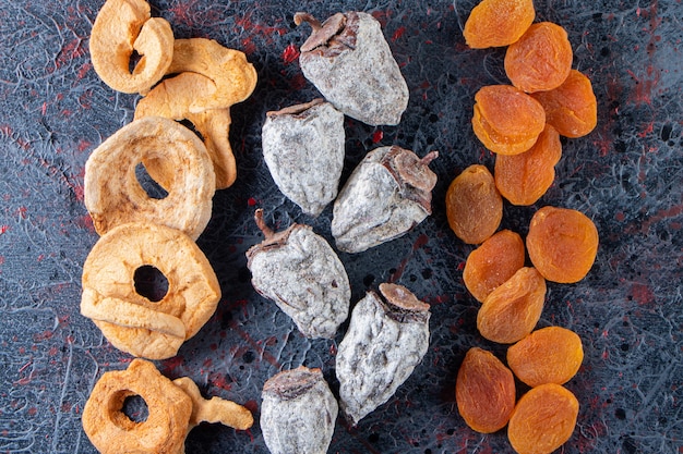 Anneaux de pomme séchés, abricots et kakis savoureux sur une surface sombre.