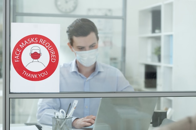 Photo annonce sur l'exigence de porter un masque de protection sur le mur de verre d'un bureau moderne avec un jeune homme d'affaires en réseau derrière