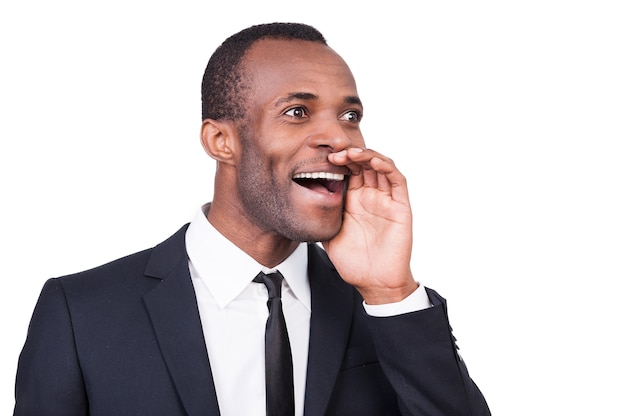 Annoncer la bonne nouvelle. Gai jeune homme africain en tenue de soirée tenant la main près de la bouche et criant en se tenant debout isolé sur fond blanc