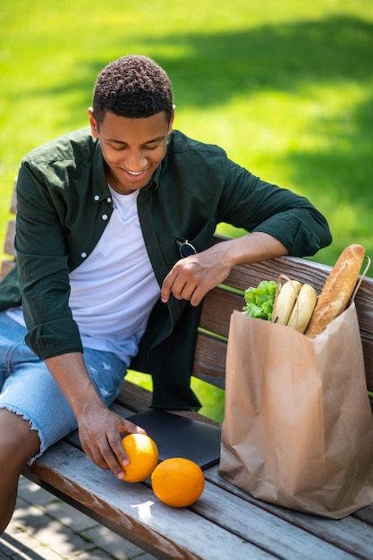 Photo anticipation. un mec souriant à la peau foncée regardant avec plaisir des oranges assis sur un banc dans le parc par beau temps