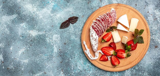 Antipasto tranché espagnol Fuet salami wurst, camembert, fraises et verre de vin rose sur fond bleu. Format de bannière longue. vue de dessus.