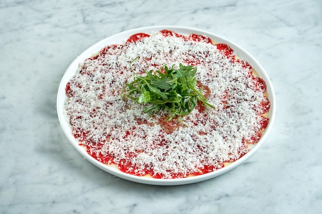 Apéritif italien classique - carpaccio de veau à la roquette et au parmesan servi sur une assiette blanche sur une table en marbre. Bœuf tranché finement.