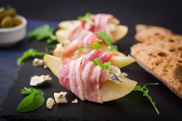 Apéritif à la poire, fromage bleu, jambon prosciutto et pain grillé pour des vacances sur une assiette sombre.