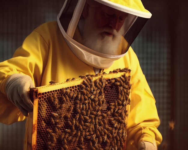 Un apiculteur en costume jaune tient une ruche avec une ruche pleine d'abeilles.