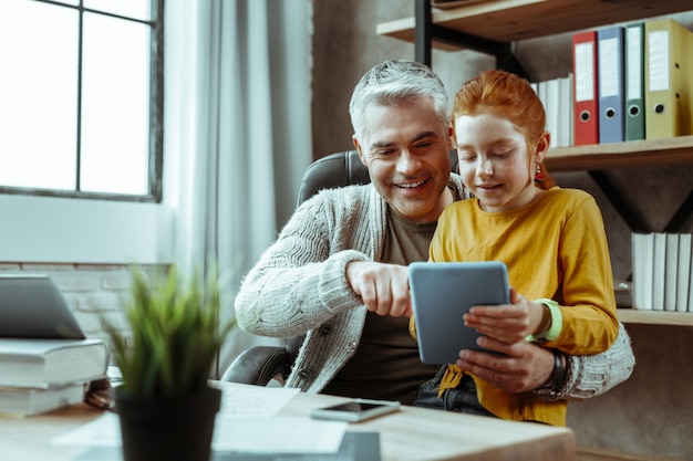 Appareil moderne. Homme positif joyeux appuyant sur l'écran de la tablette tout en montrant l'appareil à sa fille