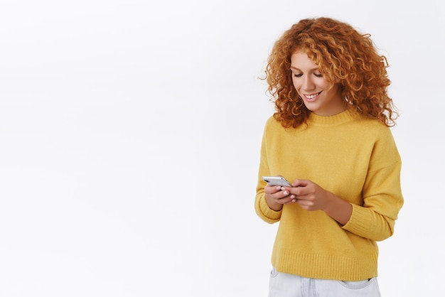 Appareils mobiles de technologie et concept de mode de vie Portrait joyeuse femme rousse bouclée en pull jaune tenant un smartphone regarder un téléphone portable avec un sourire heureux envoyer des SMS en regardant une vidéo amusante