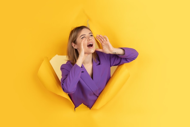 Appeler, crier. Une jeune femme caucasienne joyeuse pose sur un fond de papier jaune déchiré, émotionnel et expressif. Rupture, percée. Concept d'émotions humaines, d'expression faciale, de ventes, d'annonces.