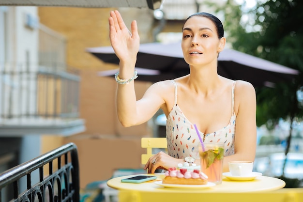 Appeler un serveur. Jolie jeune femme assise à la table à l'extérieur et mettant sa main tout en appelant un serveur